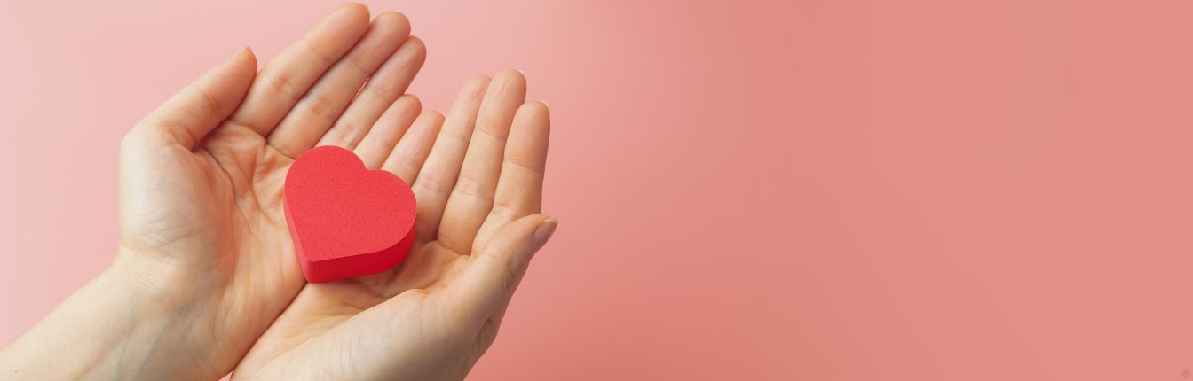 Two hands holding a red heart over a pink background.