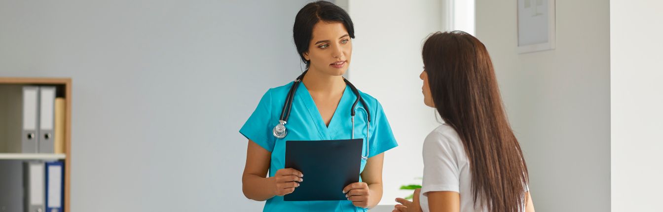 A woman with long brown hair facing away from the camera, talking to her doctor.