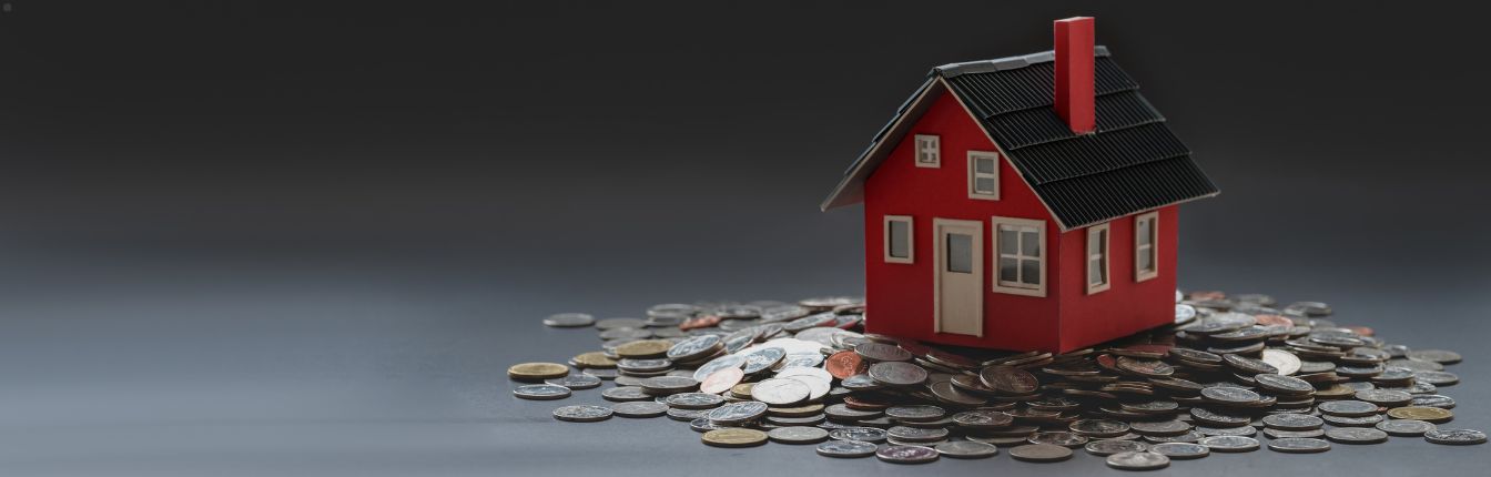 A small, red model house on top on a pile of coins, against a dark grey background.