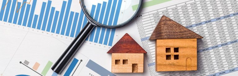 Two small wooden houses and a magnifying glass on top of papers showing financial charts and graphs.